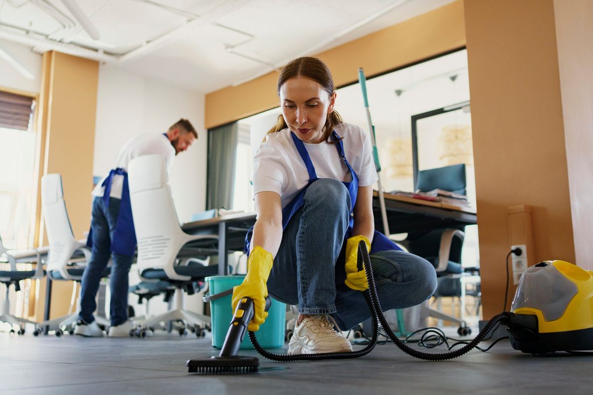 Two cleaners steam-cleaning the floor and wiping desks in an office